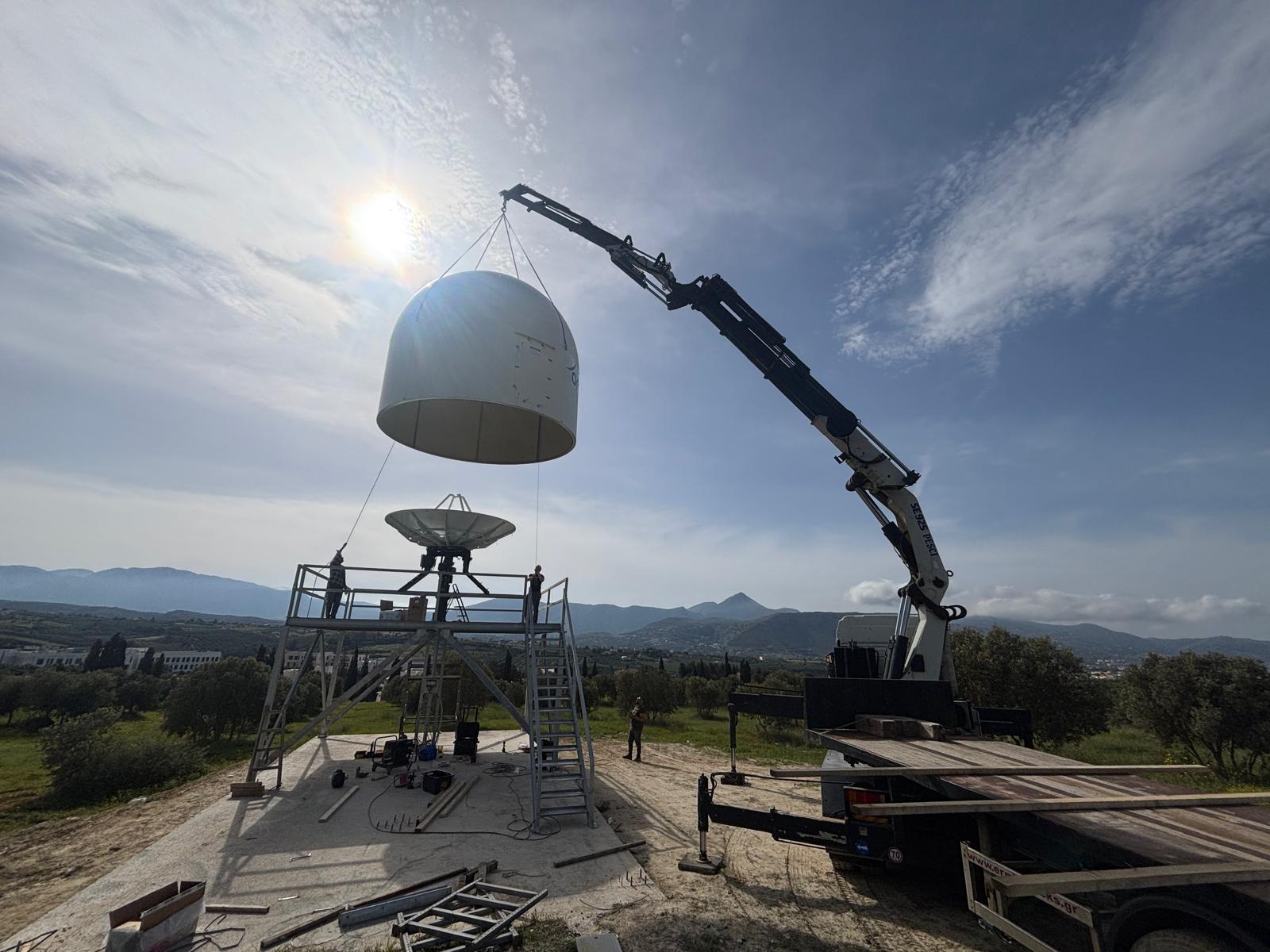 S-band ground-station dome being lifted by crane onto its antenna tower — HELEO ground-segment installation in Greece
